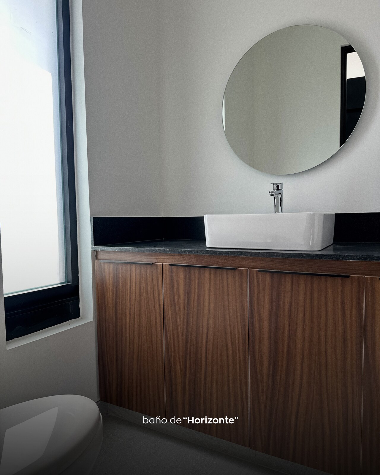 Baño de visitas con espejo redondo, lavabo blanco y cubierta de granito negro · Casa Horizonte